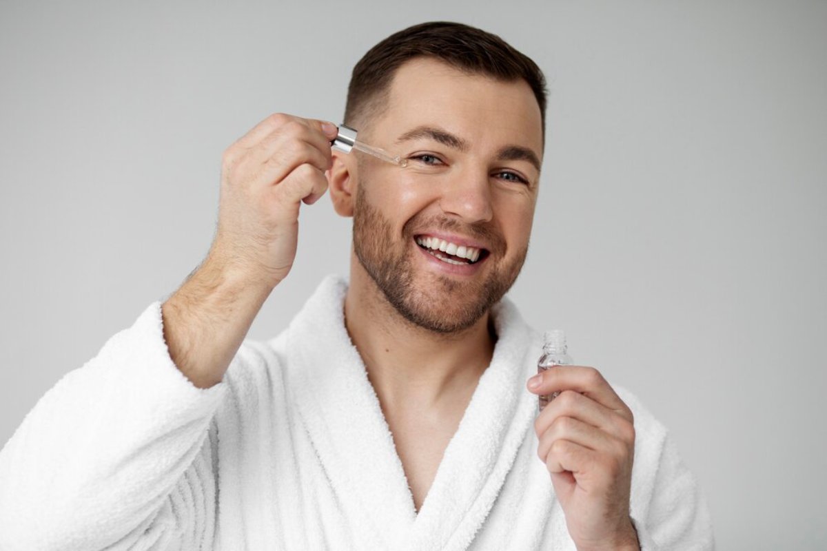 A person in a white bathrobe holds a skincare serum in one hand and a dropper in the other, against a neutral backdrop.