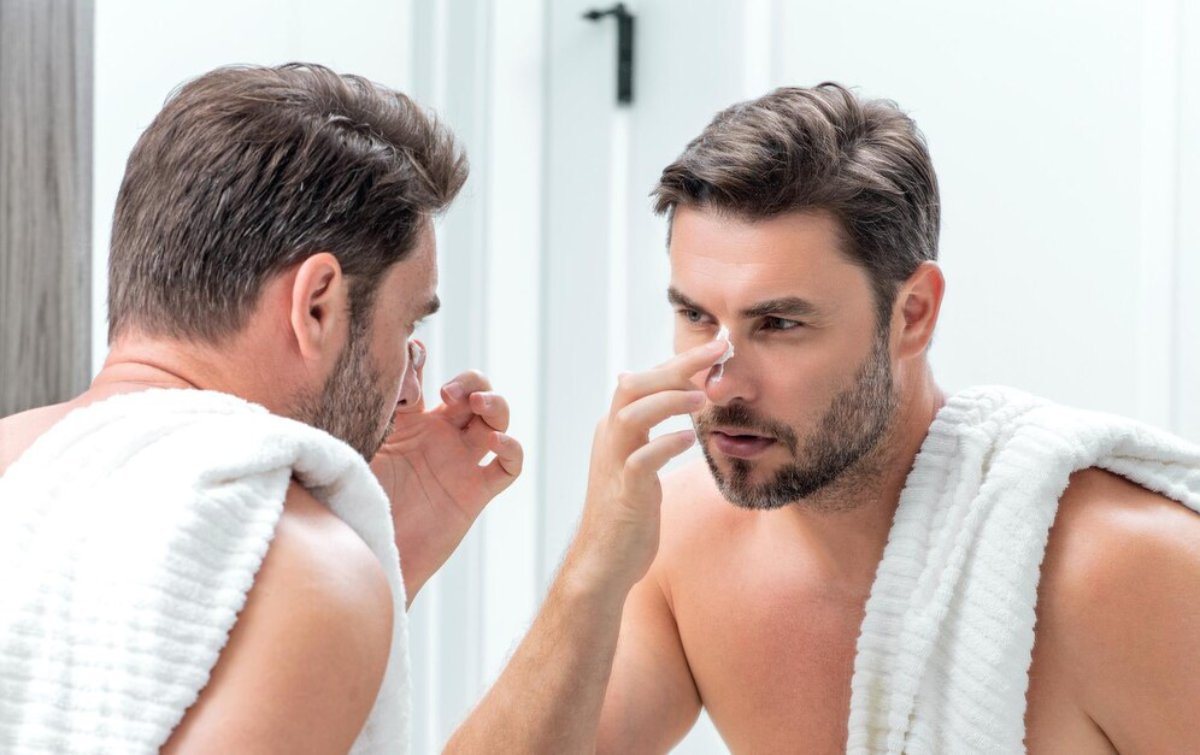 A man stands in front of a mirror, wrapped in a white towel, grooming himself in a bright, modern bathroom.
