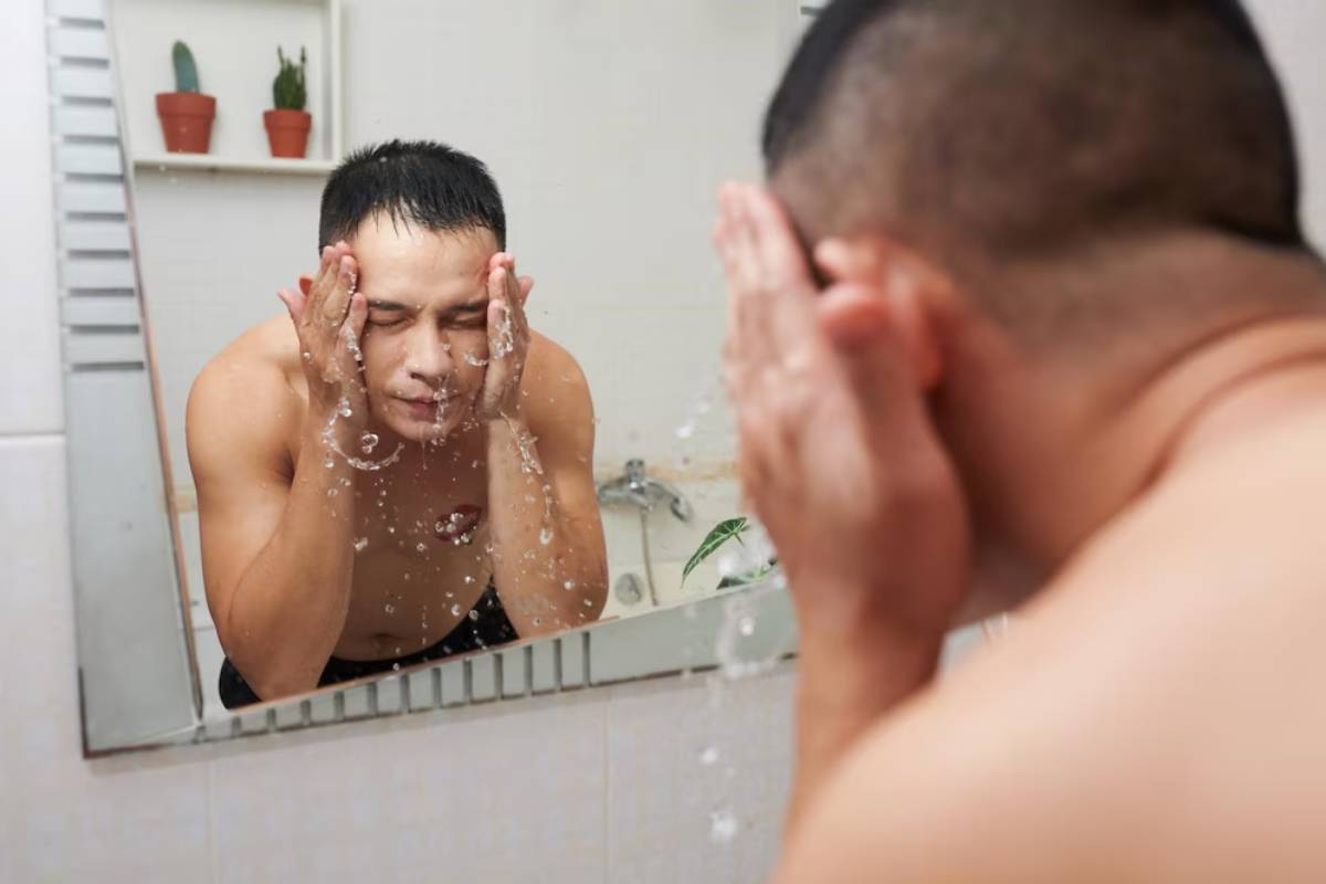 A person in a bathroom washes their face in front of a mirror, water splashes around, while potted plants sit on a shelf nearby.