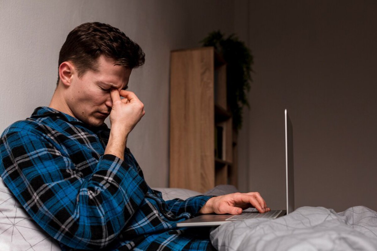 A person in a blue plaid shirt sits on a bed, typing on a laptop, with a cozy room backdrop featuring a bookshelf and a plant.