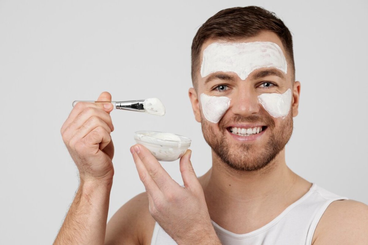 A person holds a small bowl of cream and a brush, preparing for a skincare or beauty treatment, wearing a sleeveless shirt.