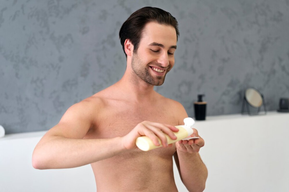 A shirtless man holds a bottle of lotion, preparing to apply it, with a minimalist background of soft textures and neutral colors.