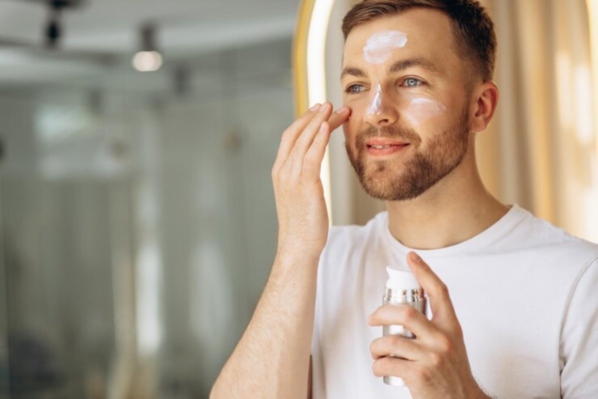 A young man in a white t-shirt applies skincare product to his face in a bright, modern bathroom with soft lighting.