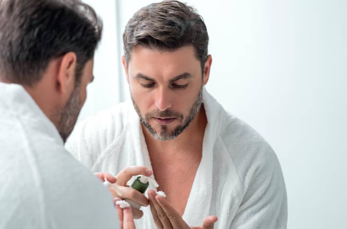 A person in a white bathrobe applies skincare products while looking in a mirror, set in a bright, minimalist bathroom.