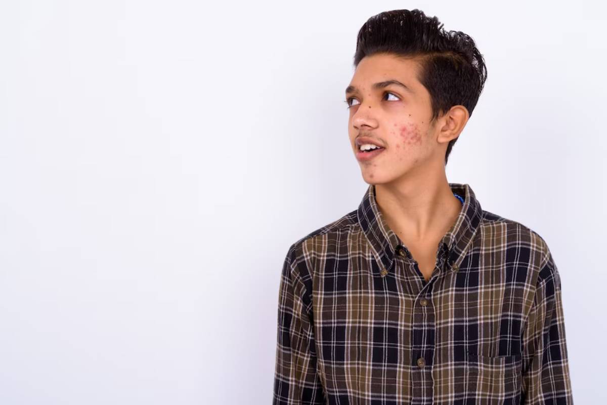 A person stands against a plain white background, wearing a checkered brown and black shirt, with a neutral expression.