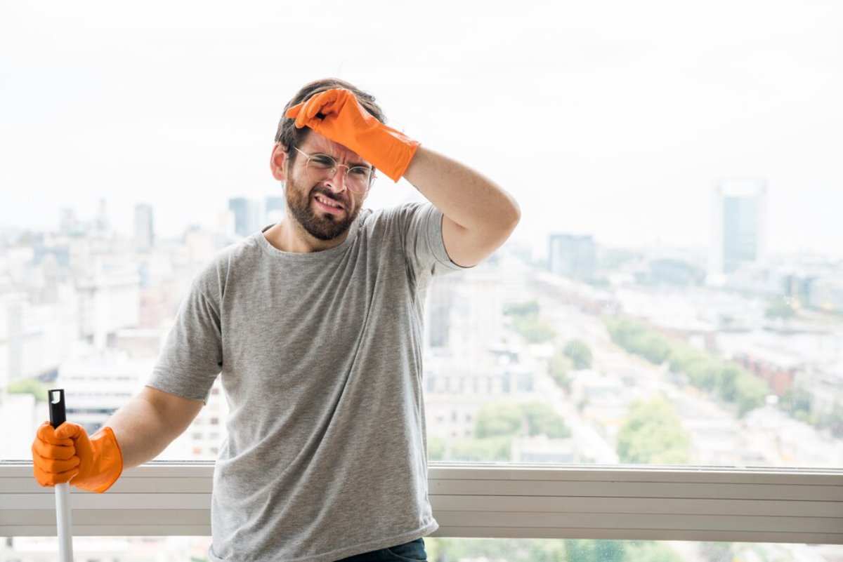 A person in a gray t-shirt and orange gloves stands by a window, holding a cleaning tool with a city skyline in the background.