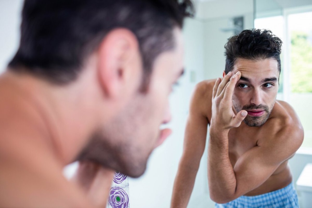 A man in a light blue checkered outfit examines himself in a mirror, showing concern while touching his face in a bright bathroom.