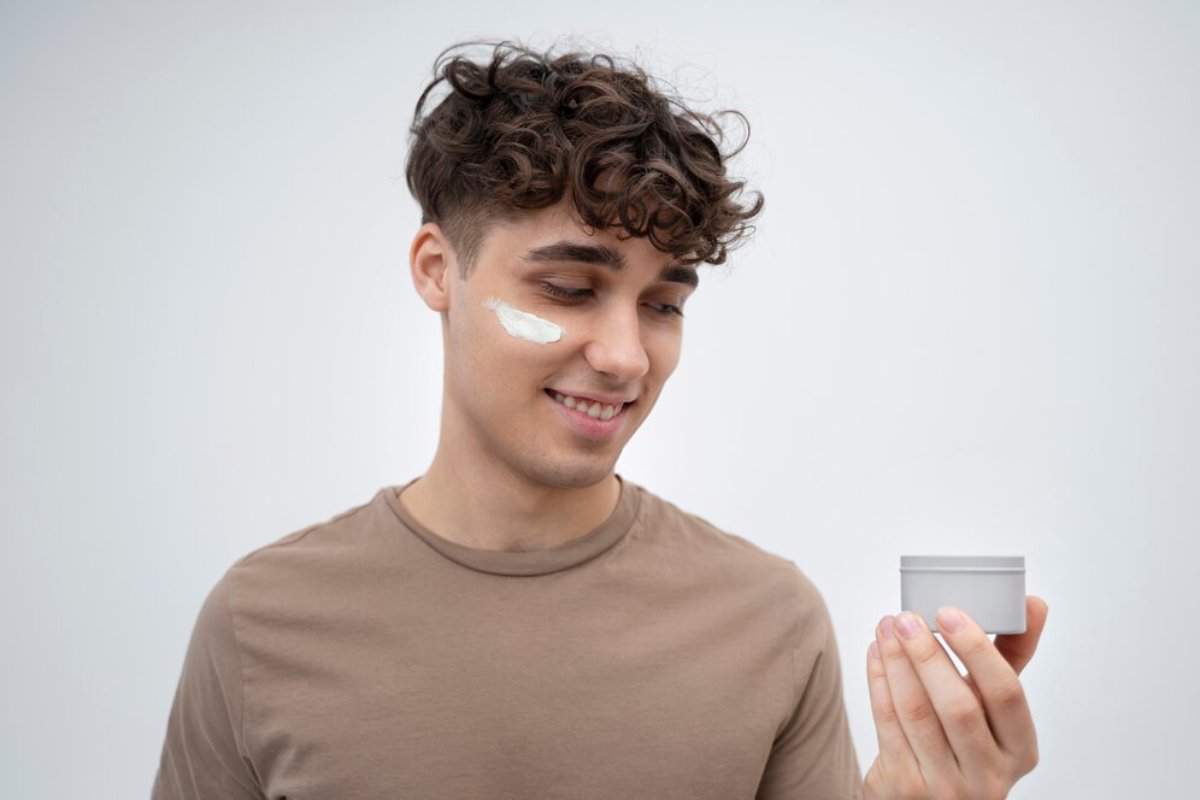 A young man with curly hair holds a small, round gray container in his right hand against a light gray background.