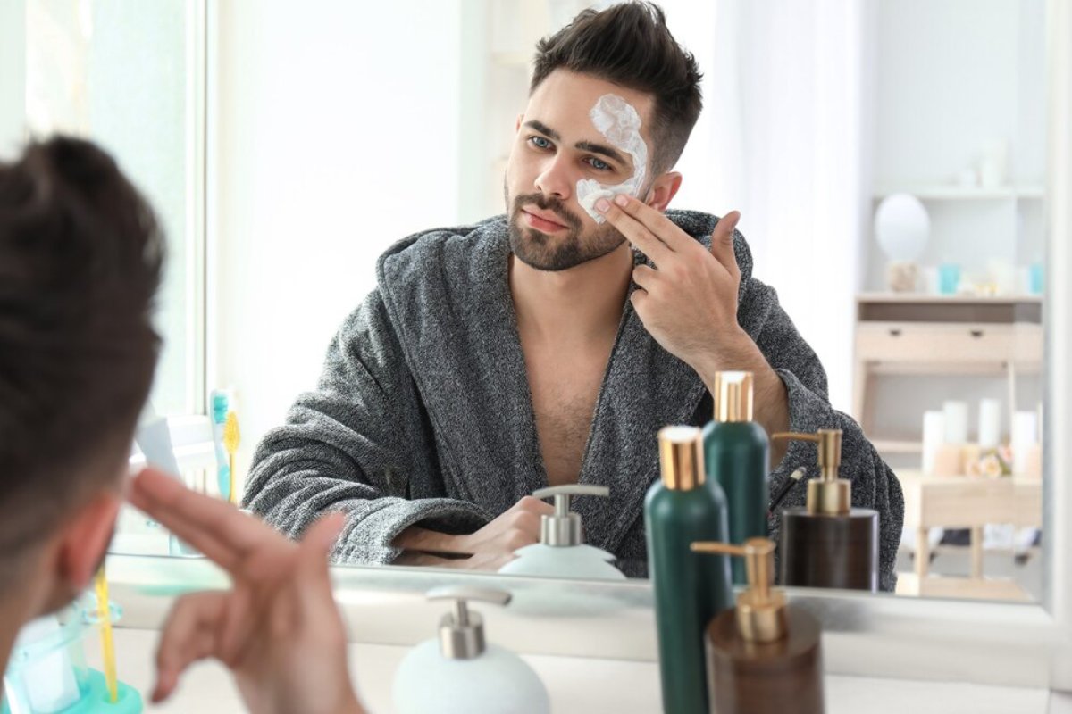 A person in a gray robe applies skincare products in a bright bathroom with various bottles and a mirror on the countertop.