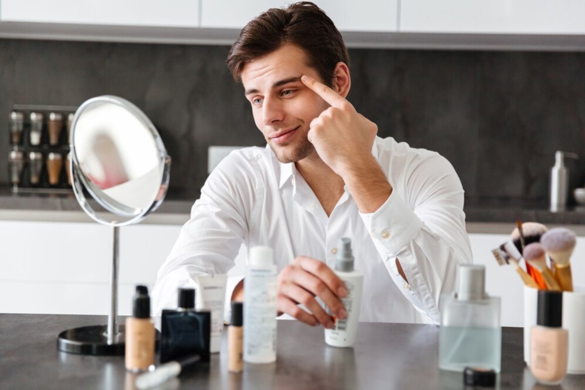 A person in a white shirt sits at a vanity, surrounded by various skincare and makeup products on a dark countertop.