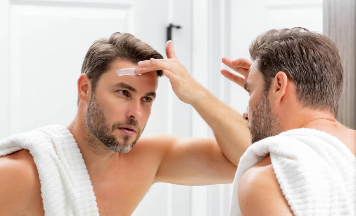 A man in a bathroom wearing a white towel inspects his hair in the mirror, focusing on styling. Bright, modern surroundings.