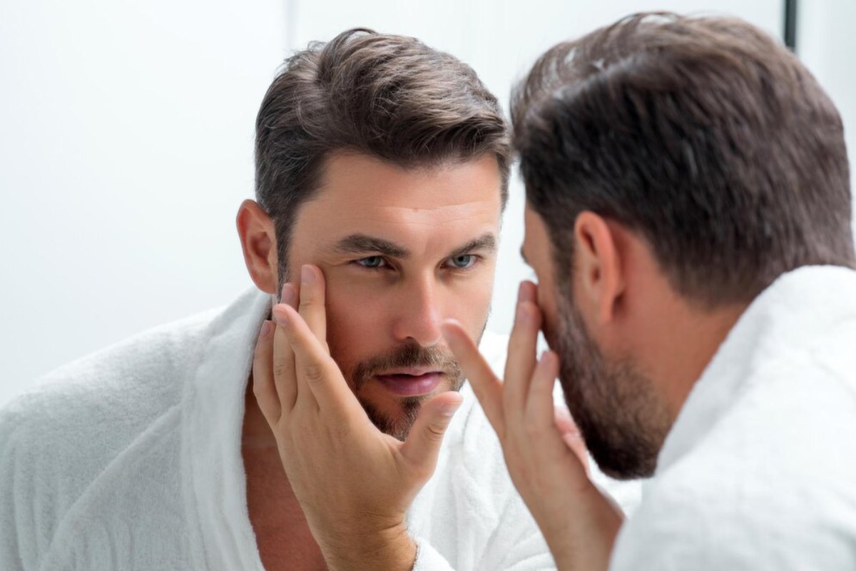 A man in a white robe examines his reflection in a mirror, contemplating as he gently touches his chin in a bright, modern bathroom.