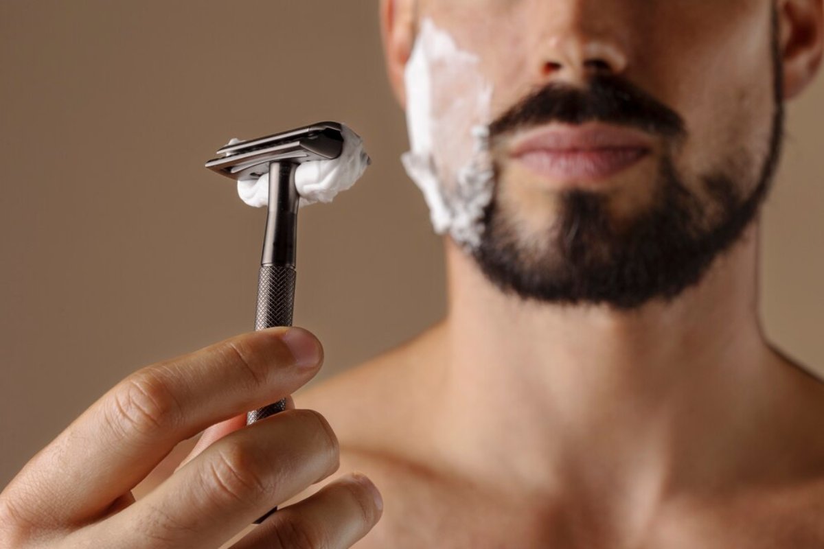 A person holding a metal razor with shaving cream on the blade, set against a neutral background.