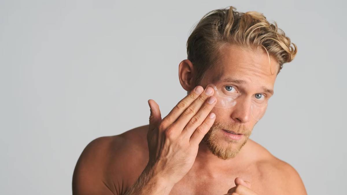 A muscular young man with wavy blonde hair gestures thoughtfully, showcasing a confident pose against a neutral background.