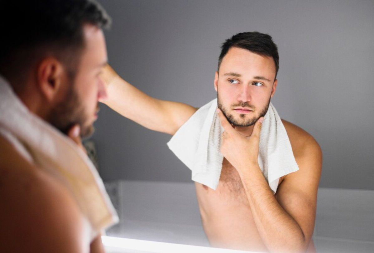 A man stands shirtless in front of a mirror, thoughtfully touching his chin while draped in a towel, with soft lighting around him.