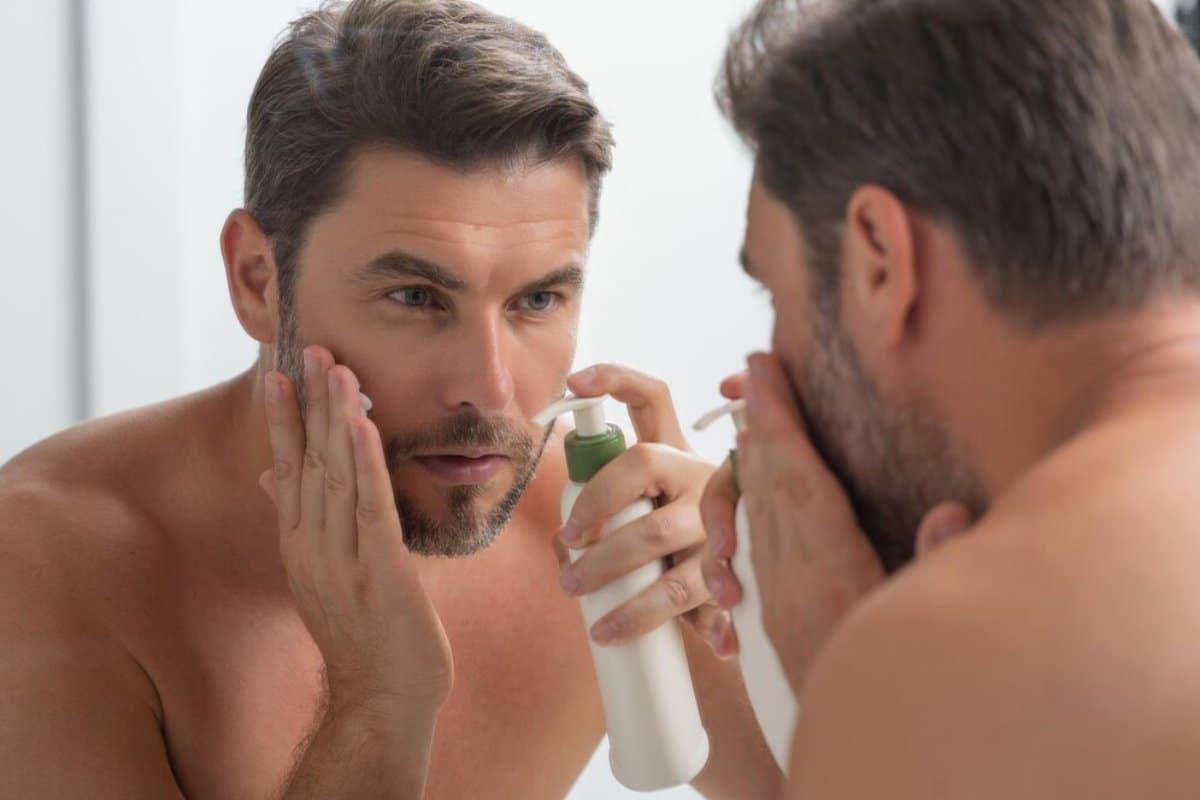 A man in a bathroom mirrors his actions while applying skincare products, emphasizing a morning routine of self-care.