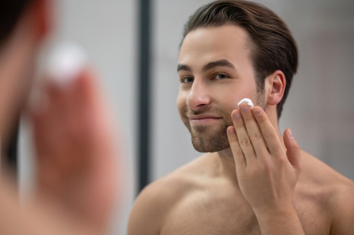 A shirtless man applies cream to his face while looking in a mirror, focusing on skincare in a modern bathroom setting.