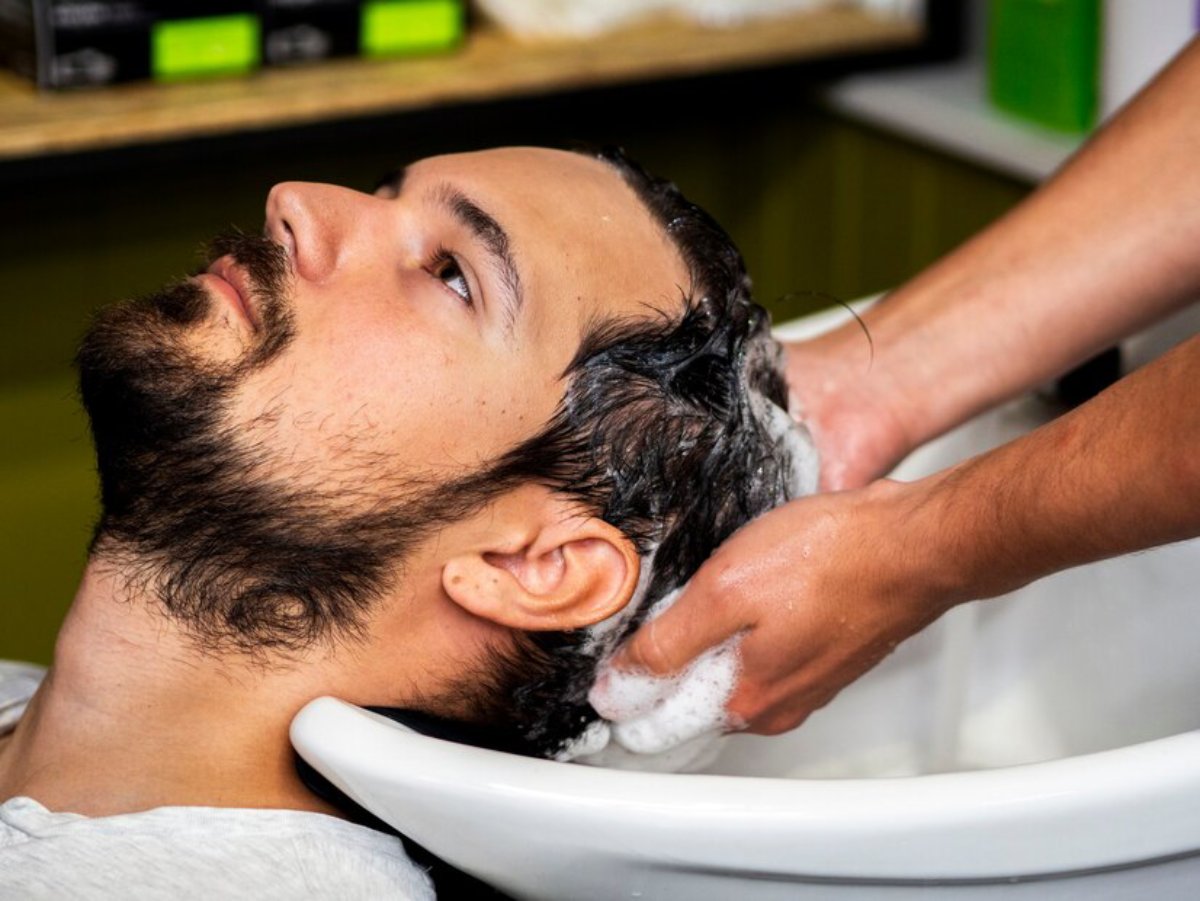 A person reclines at a salon sink while receiving a hair wash, with soothing bubbles lathered on their hair by a stylist.