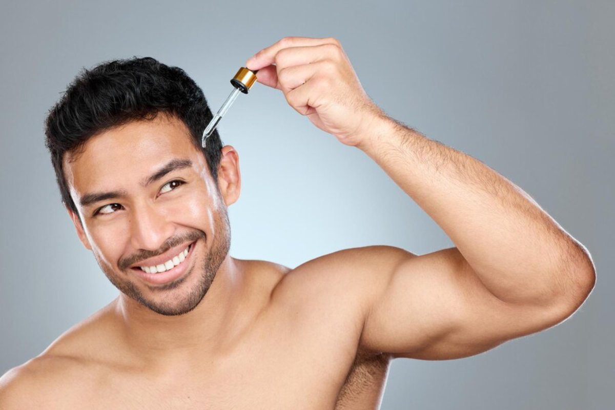 A person with dark hair holds a dropper above their head, preparing to apply a liquid product, against a soft gray background.