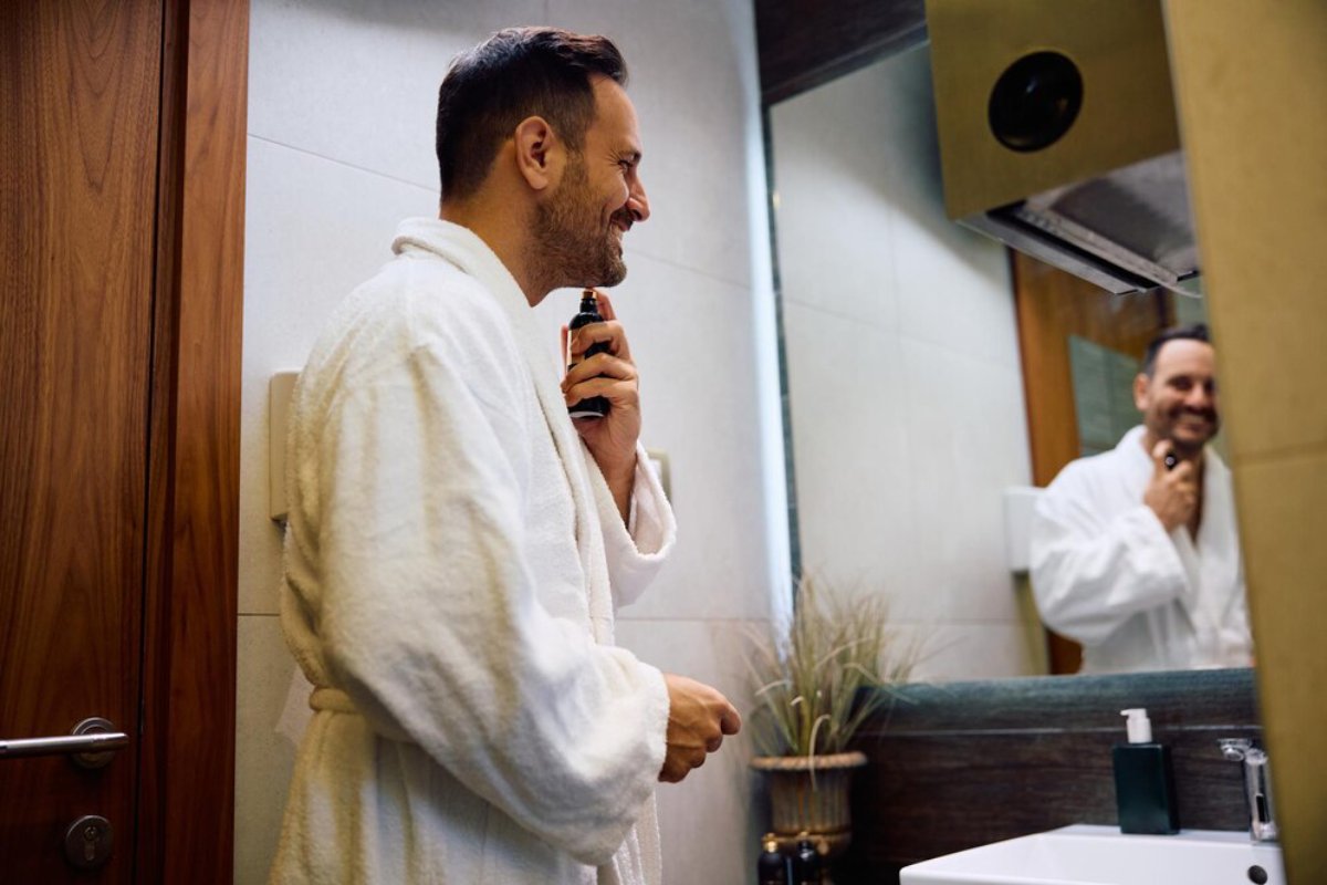 A man in a white bathrobe stands in a bathroom, holding a grooming spray while looking into a mirror.