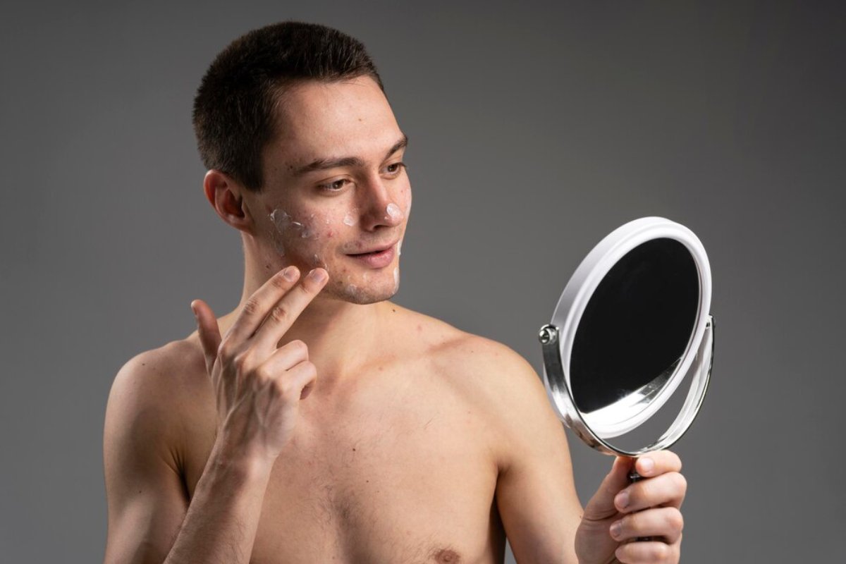 A shirtless young man holds a handheld mirror, examining his reflection with a thoughtful expression. Soft lighting highlights his features.