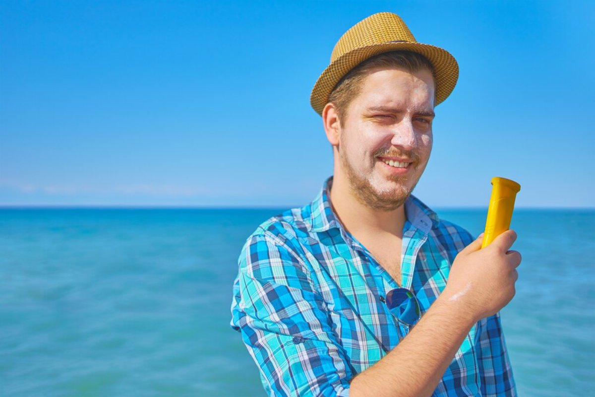 A person in a straw hat and a blue plaid shirt stands by the sea, holding a yellow ice cream cone against a bright blue sky.
