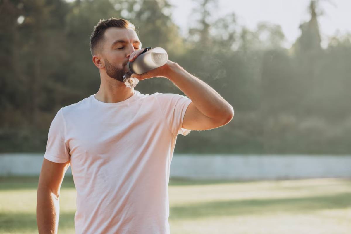 A man in a white t-shirt drinks from a water bottle while standing on a grassy field, surrounded by trees in soft sunlight.