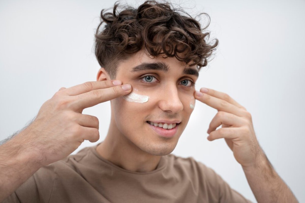 A person with curly hair is pointing at their face with both hands, wearing a light brown shirt against a neutral background.
