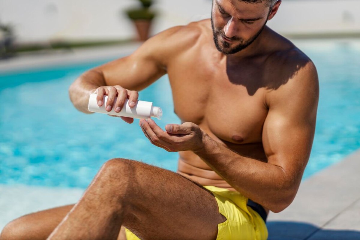 A man sitting by a pool applying sunscreen lotion to his hand, wearing yellow swim trunks, with a sunny day in the background.