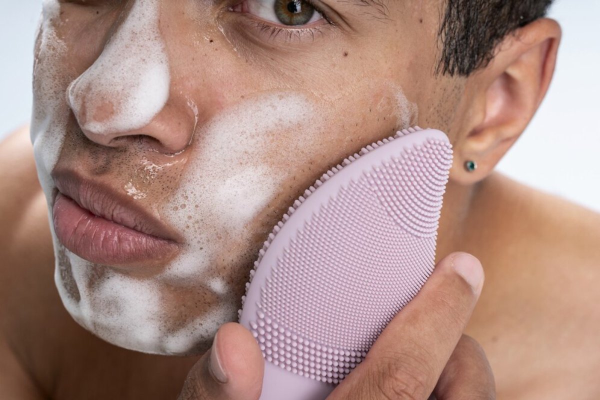 A close-up of a person holding a facial cleansing brush, showcasing their ear with a subtle earring and part of their shoulder.