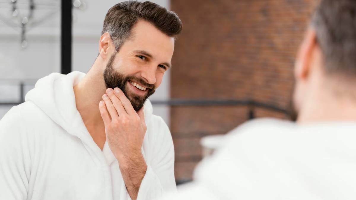 A man in a white bathrobe touches his chin while looking into a mirror in a modern, stylish interior setting.