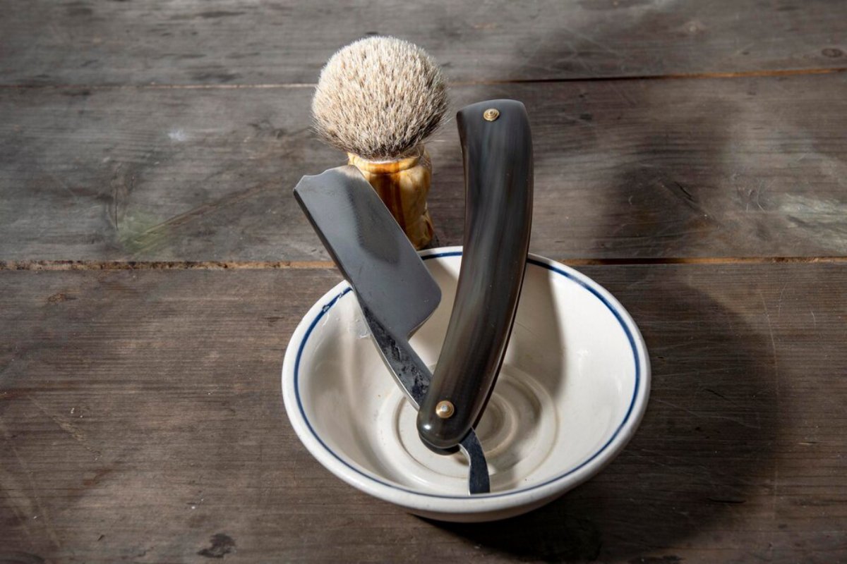A straight razor rests in a shallow bowl beside a soft-bristled shaving brush, all set on a rustic wooden surface.