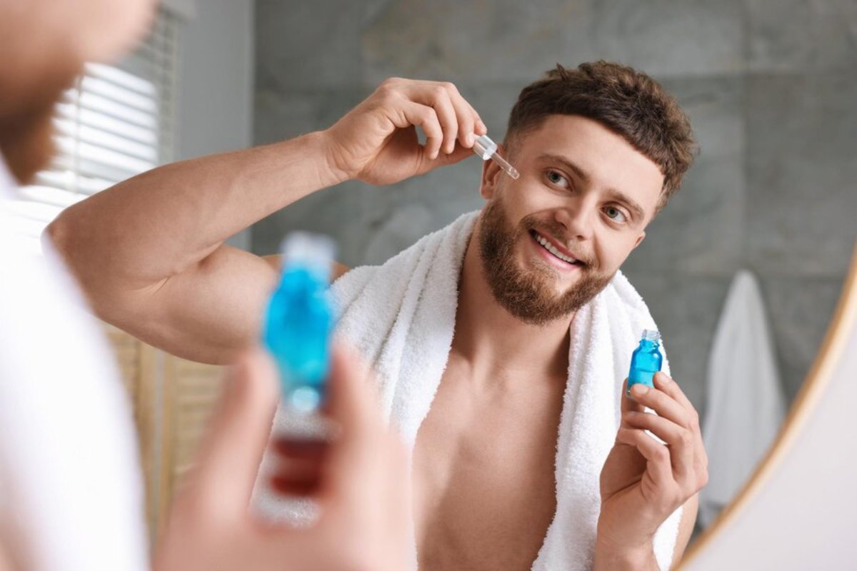 A man in a towel holds a small blue bottle while inspecting himself in the bathroom mirror, showcasing a grooming routine.