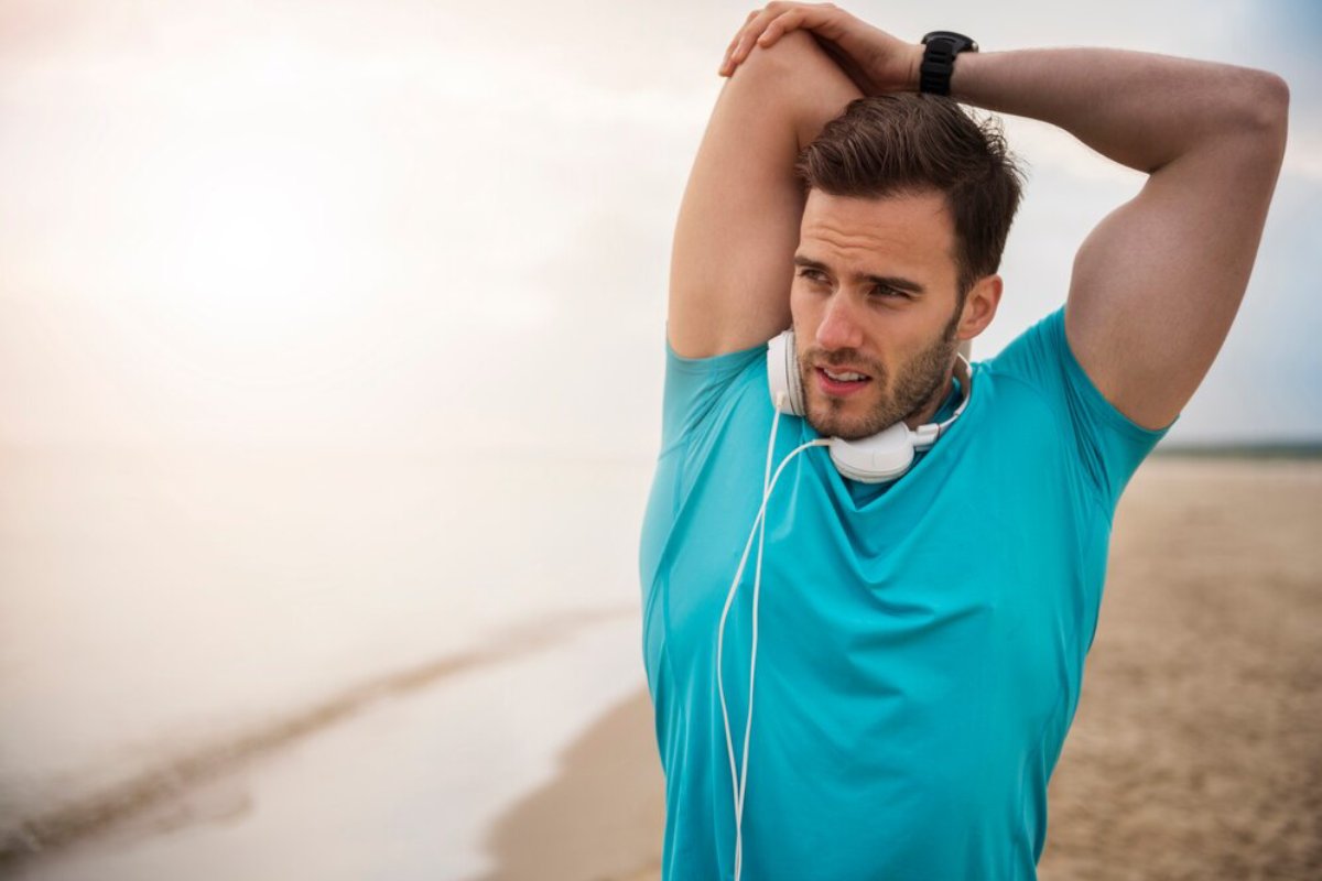 A person wearing a blue athletic shirt stretches on a beach, with headphones around their neck, as the sun sets in the background.