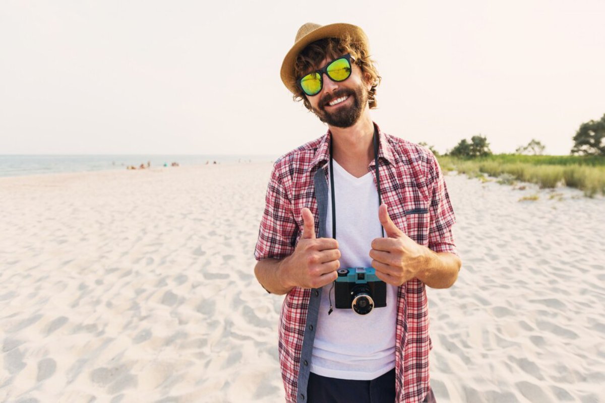 A person stands on a sandy beach, wearing a plaid shirt, cream hat, and holding a camera, displaying thumbs-up in a joyful pose.