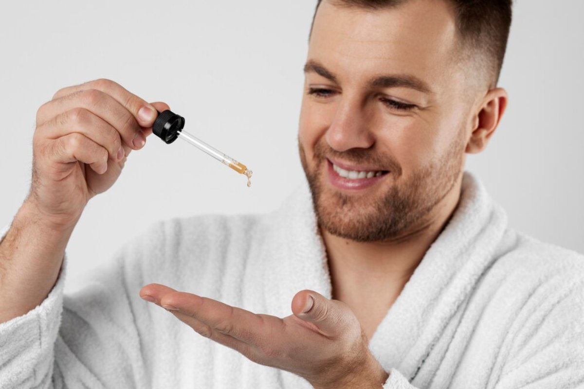 A person in a white robe holds a dropper above their palm, ready to apply a skin serum against a plain white background.