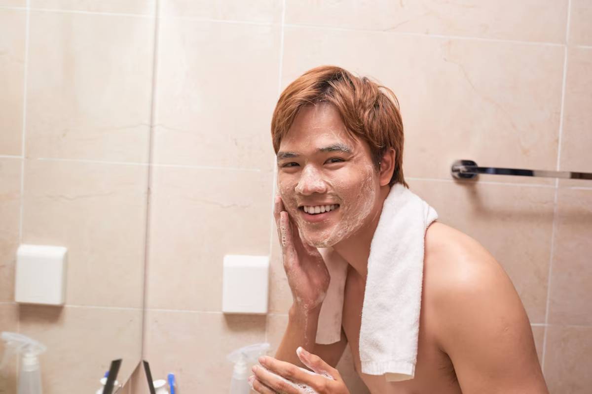 A person with short, light brown hair is washing their face at a bathroom sink, wearing a towel around their neck.