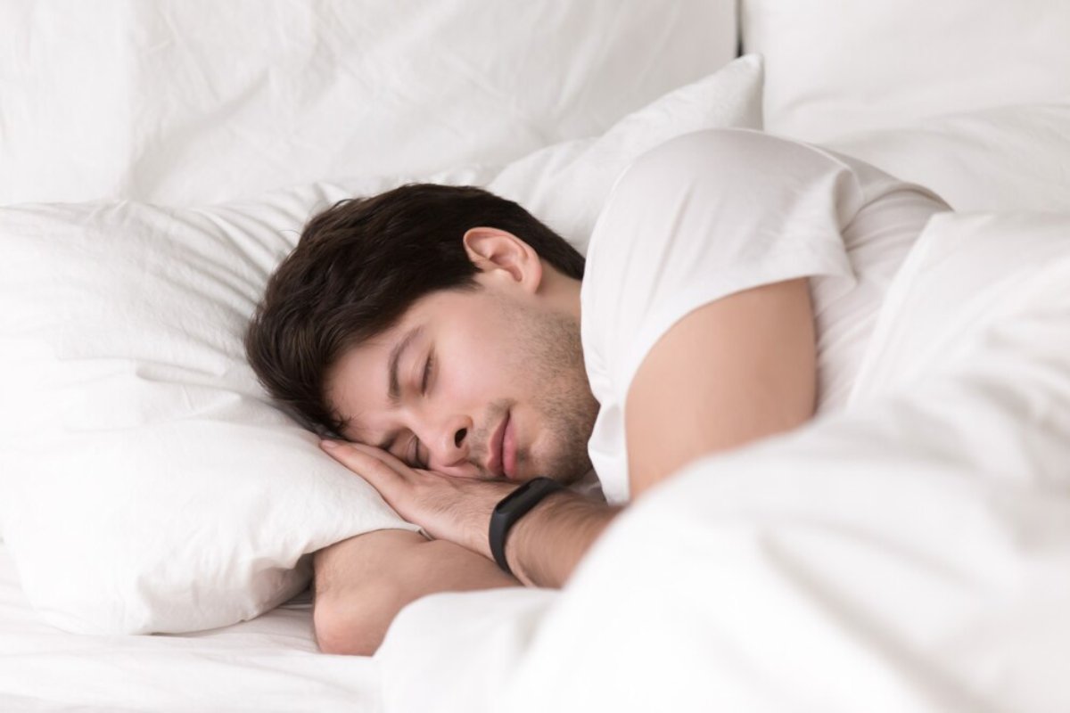A person lies curled up in bed, surrounded by fluffy white pillows, wearing a simple white t-shirt, appearing peaceful and relaxed.