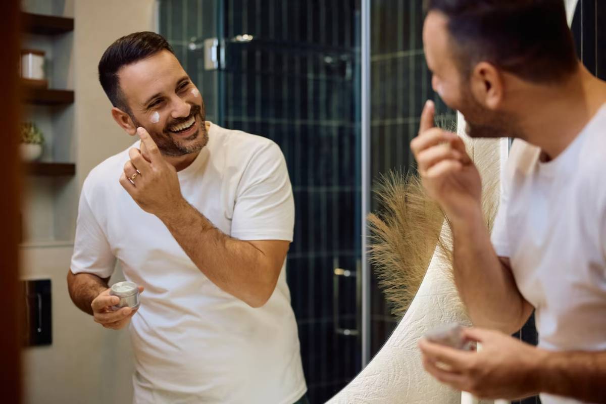 A man in a white t-shirt applies skincare product while looking in a bathroom mirror, surrounded by modern decor.