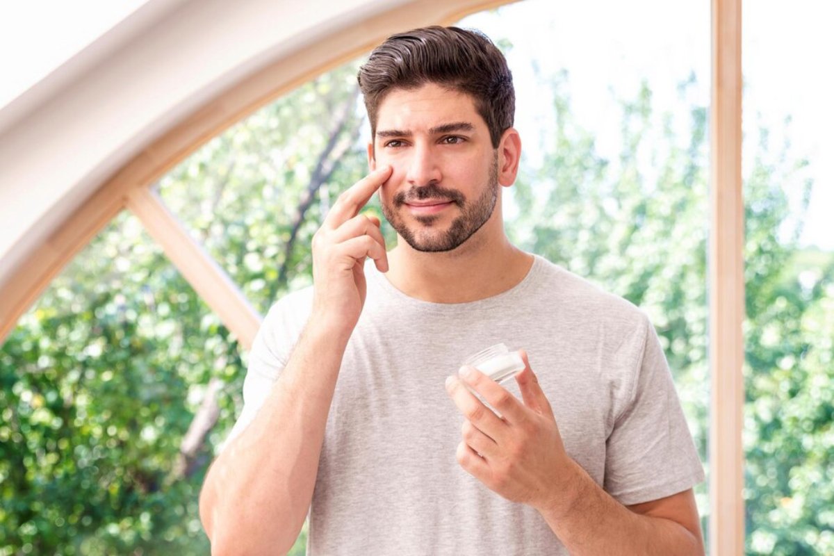 A person in a gray t-shirt applies skincare product while holding a jar, standing in a bright room with greenery outside.