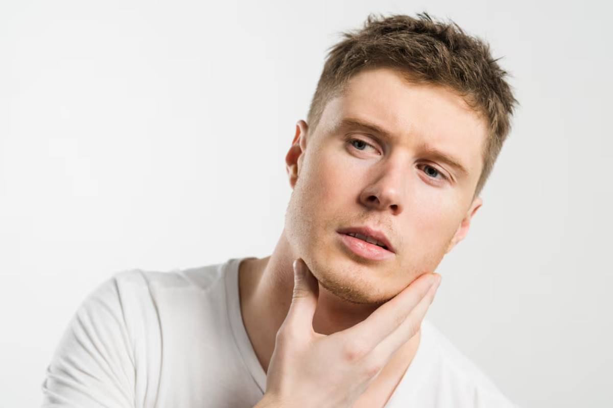 A young man in a white t-shirt is holding his chin thoughtfully, set against a bright, neutral background.