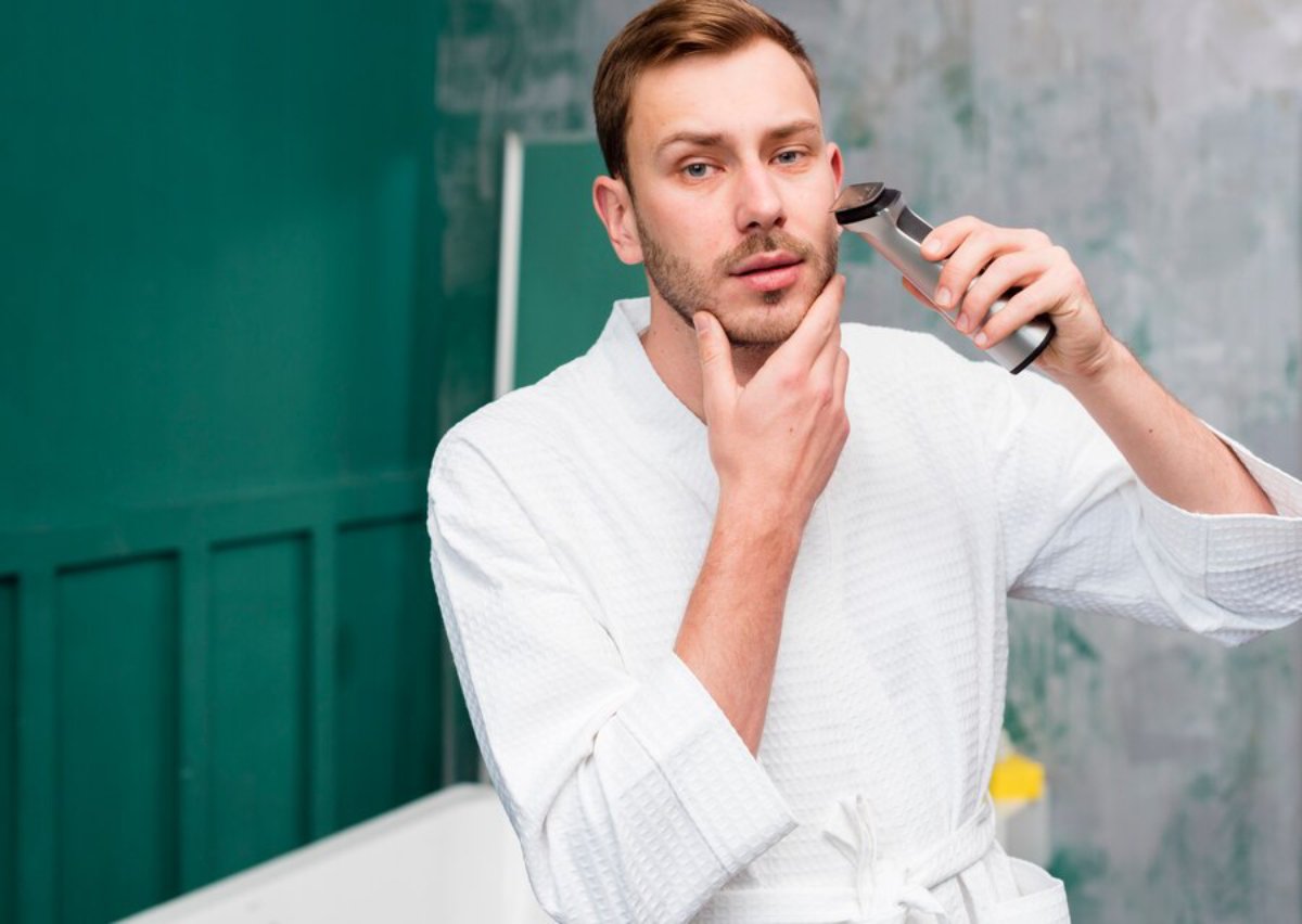 A person in a white robe uses an electric trimmer while standing in a modern bathroom with green walls.
