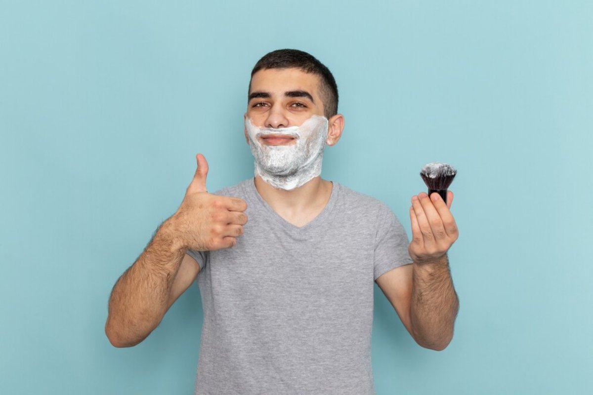 A person in a gray t-shirt gives a thumbs up while holding a shaving brush, with a light blue background.