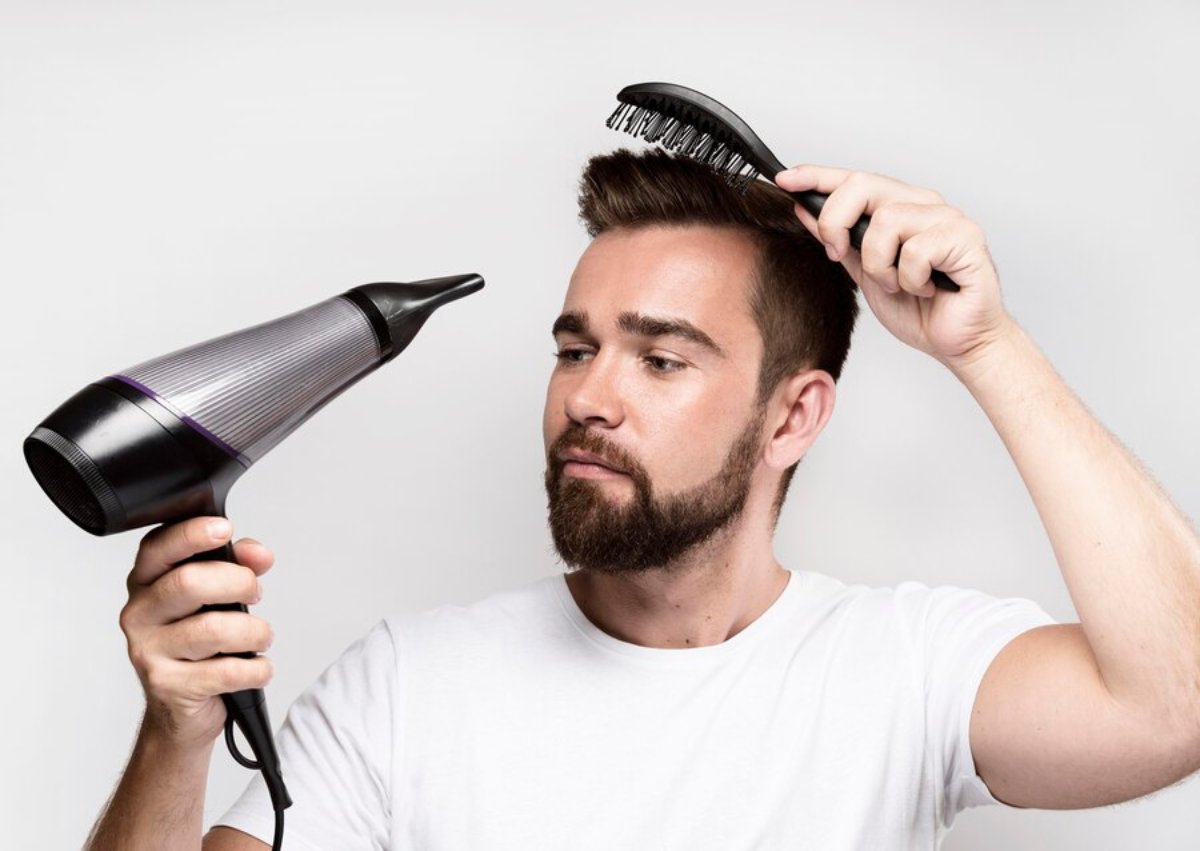 A person in a white shirt holds a hairdryer in one hand and a brush in the other, styling hair against a plain background.