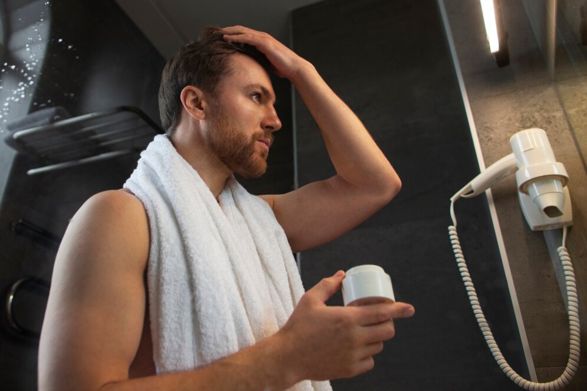 A man in a white towel stands in a bathroom, holding a container and running his hand through his hair in a mirror-lit space.