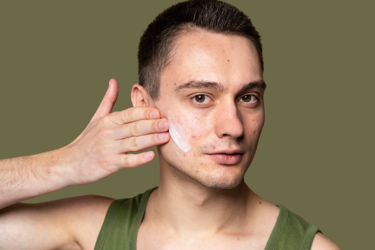 A person in a green tank top holds their hand to their ear, against a solid olive green backdrop.