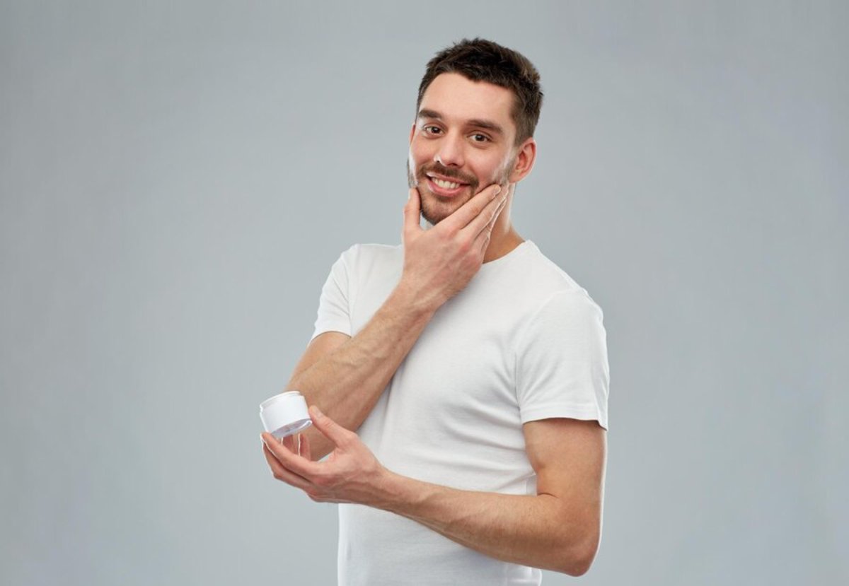 A man in a white t-shirt holds a jar of cream while contemplating, against a simple gray background.