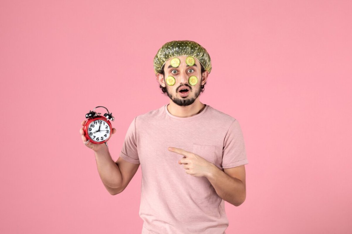 A person in a light pink shirt holds a vintage red alarm clock, pointing at it against a pastel pink background.