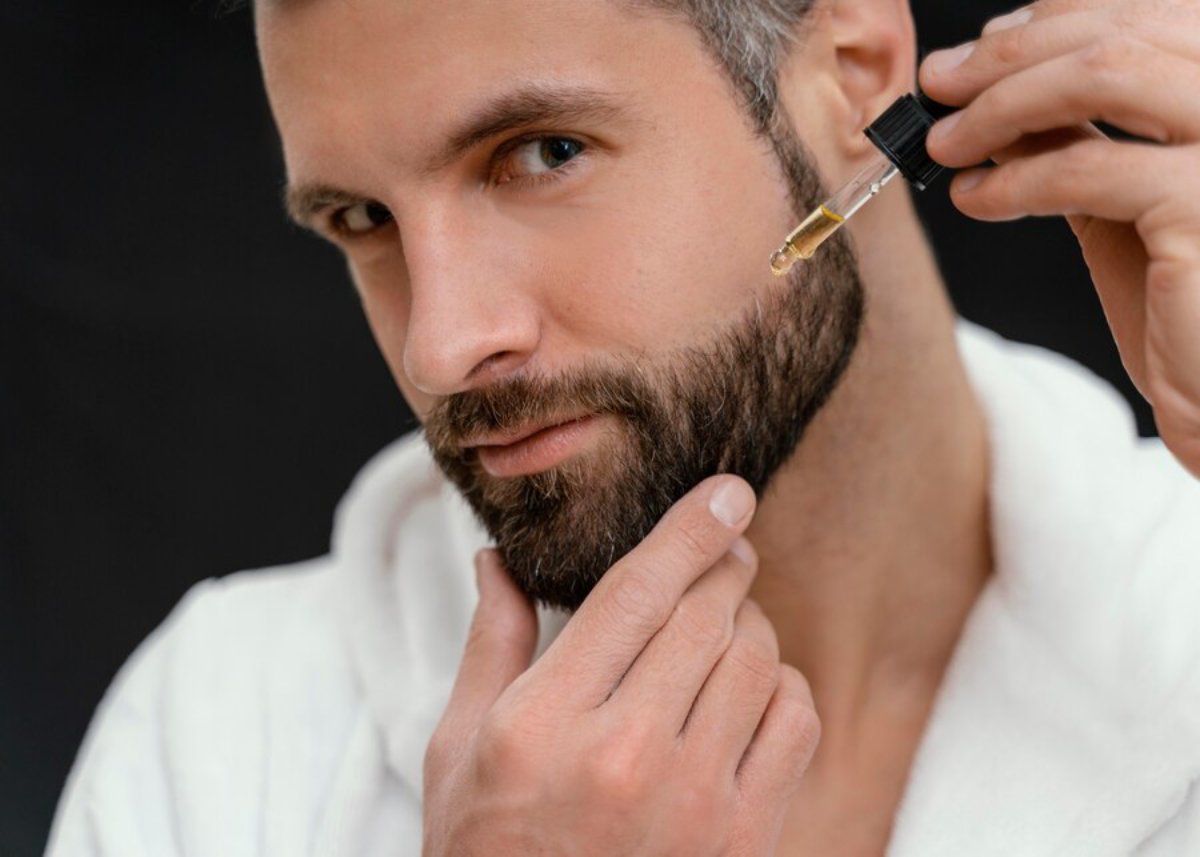 A person in a bathrobe applies beard oil using a dropper, focusing on their grooming routine against a dark background.