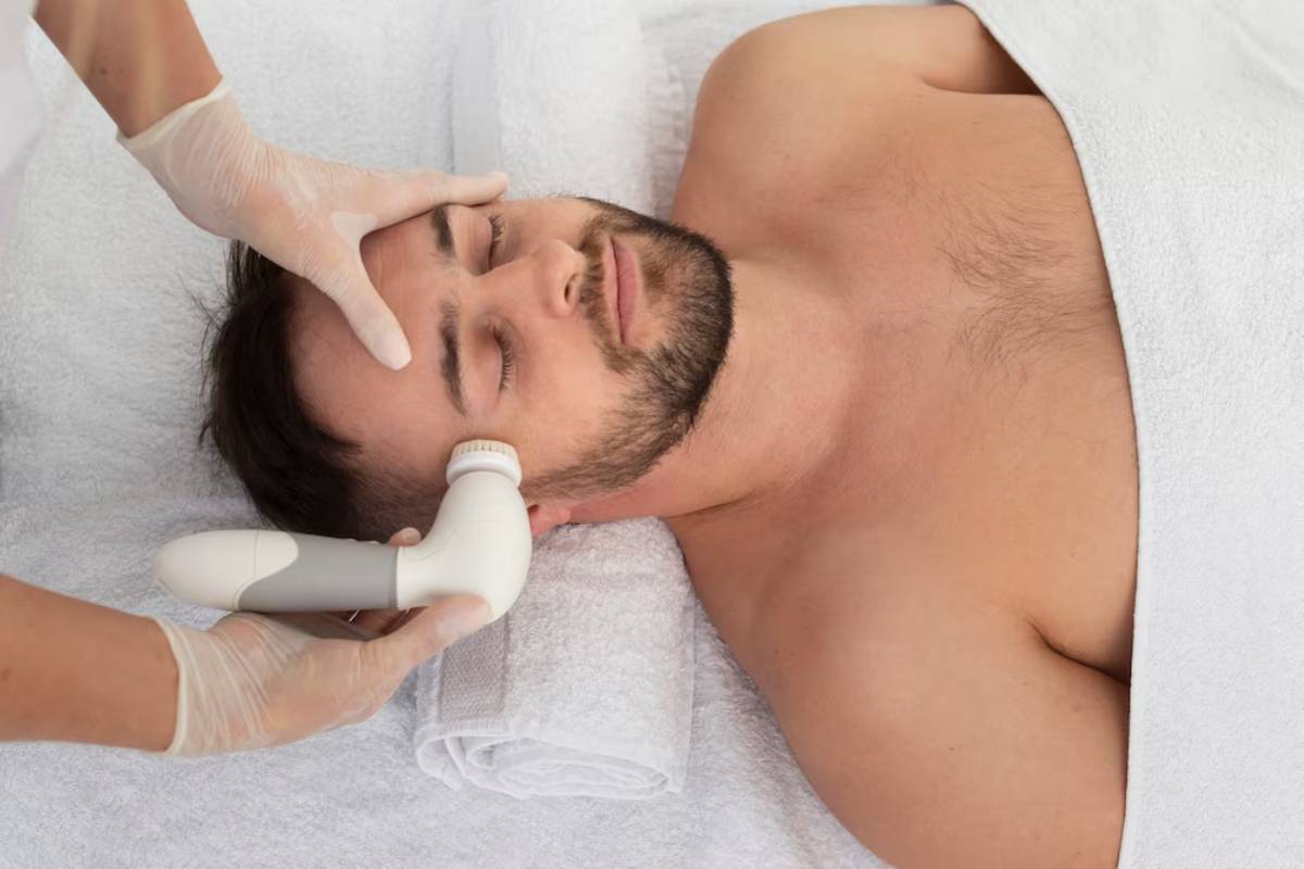 A person receives a facial treatment with a handheld device, their torso covered in a white towel, hands protected with gloves.